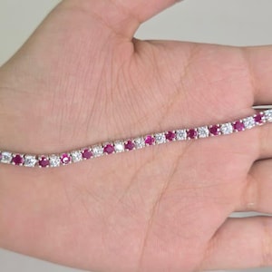 May include: A silver-coloured tennis bracelet with alternating red and clear gemstones. The bracelet is displayed on a hand.