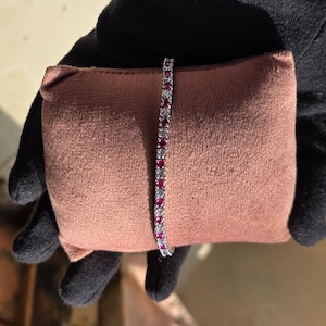 May include: A tennis bracelet featuring a repeating pattern of round, deep red gemstones alternating with clear, sparkling stones set in a polished silver-toned metal band. The bracelet is displayed vertically against a soft, rectangular cushion.