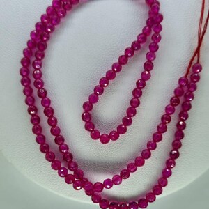 Pode incluir: Um colar ou pulseira feito de pequenas pedras preciosas rosa-escuras facetadas. As contas são redondas e polidas, refletindo a luz. O fundo é uma superfície branca lisa. As pedras preciosas são provavelmente contas.