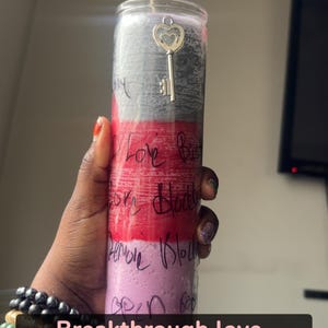 Puede incluir: Una vela cilíndrica alta en un recipiente de vidrio transparente con bandas horizontales de cera gris, roja y rosa claro. La vela tiene un dije de llave en forma de corazón de tono plateado cerca de la parte superior y texto negro escrito.