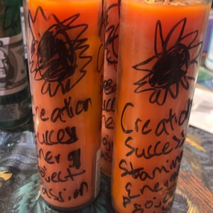 May include: Three orange glass candle jars with black handwritten text on the front. The text reads "Creation Success" and "Stamina Energy Passion".