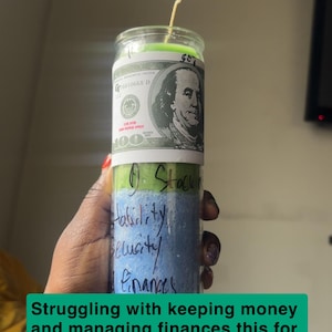 May include: A cylindrical glass candle with a green wax top and blue wax base. The label features a 100 dollar bill graphic with Benjamin Franklin. Handwritten text on the glass reads D Stack n save, Stability, Security, and Finances.