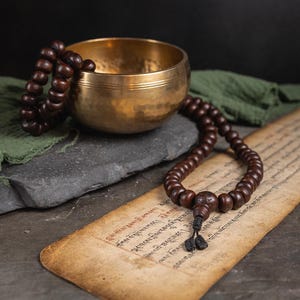 May include: A golden singing bowl with a brown wooden mala bead necklace resting on top of it. The bowl is sitting on a dark grey stone. The mala beads are draped over a piece of green fabric and an old, yellowed piece of paper with writing in a foreign language.