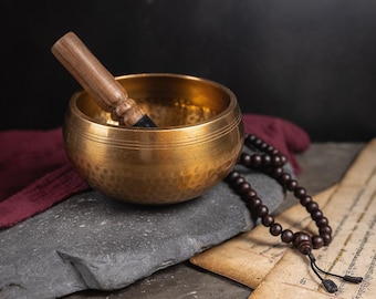 Puede incluir: Un cuenco cantor dorado con un mazo de madera, un collar de cuentas mala marrón oscuro y un trozo de pergamino con texto en un idioma extranjero.