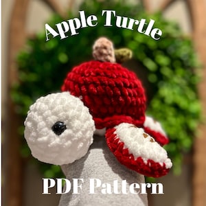 Può includere: Una tartaruga a forma di mela all'uncinetto fatta a mano con corpo rosso, testa e piedi bianchi e gambo verde. Il testo "Apple Turtle" e "PDF Pattern" è visibile nell'immagine. La tartaruga è su uno sfondo verde sfocato.