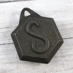 May include: A dark gray, hexagonal pendant with a loop at the top. The pendant features the letter "S" in a stylized font. The pendant is made of metal and has a textured surface.