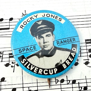 May include: A vintage, round button with a light blue background featuring a portrait of a man in a space ranger uniform. The button reads "Rocky Jones Space Ranger" and "Silvercup Bread."
