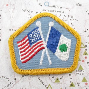 May include: A pentagonal embroidered patch with a light blue background and a thick gold border. The patch features two crossed white flagpoles with an American flag on the left and a blue and white flag with a green clover on the right.