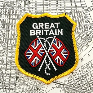 May include: A black and yellow embroidered patch with the words "GREAT BRITAIN" and two crossed Union Jack flags. The patch is set against a vintage map background.