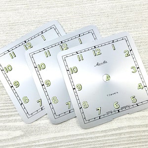 May include: Three square watch faces with a silver finish and black borders. The faces feature black minute markers, green numerals, and the brand name "Manthe". The text "7 Jewels" and "Made in Germany" are also visible.