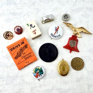 May include: A collection of vintage pins, tokens, and charms including an orange Drive-In sign, a mahjong tile, a gold eagle bell pin, a black circular token, a brass locket, a Bingo token, and several round buttons with various graphic designs.