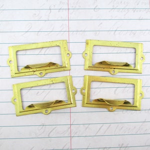 May include: Four vintage brass label holders. Each rectangular frame has a curved handle and two small holes for mounting. The brass has a slightly aged, tarnished appearance, suggesting they are antique or vintage.