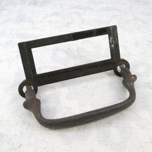 May include: A dark, weathered metal drawer pull featuring a rectangular label holder frame. The hardware includes a curved handle attached to two circular mounting points with visible threaded screws, showcasing a distressed, aged finish throughout.