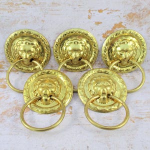 May include: Five gold-colored lion head drawer pulls with ring handles. The pulls have a textured finish and are arranged on a light-colored wooden surface. Each pull features a detailed lion's face.