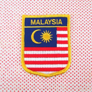 May include: Embroidered patch featuring the flag of Malaysia. The patch has a blue field with a yellow crescent and sun, and red and white stripes. The word "MALAYSIA" is embroidered in yellow at the top, with a gold border.