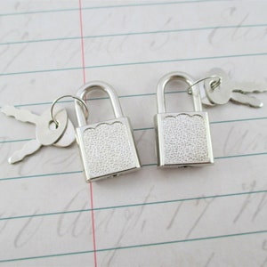May include: Two small, silver padlocks with matching keys. The padlocks have a textured, rectangular face and a curved handle. The keys are flat and have a simple, symmetrical design. The items are on a lined paper background.