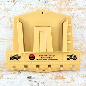 May include: A beige plastic wall-mounted organizer featuring a central mail slot and six hooks for keys. The front displays black graphics of keys and text reading Steuben County Farm Bureau Co-op The Co-op - Helping You Help Yourself.