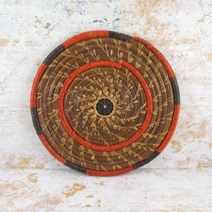 May include: A round, woven basket with a spiral design. The basket features concentric circles of brown, orange, and black. The center is a dark brown circle. This decorative item is suitable for home decor.