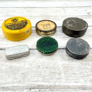 May include: A collection of vintage metal tins in various shapes and sizes. The tins are in yellow, green, silver, and dark gray. Some tins have text, including "SWARTCHILDE CO" and "BENJ ALLEN & CO". The tins are arranged on a light wood surface.