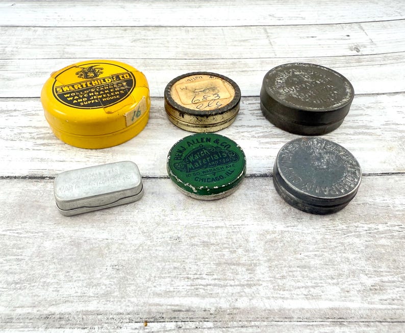 May include: A collection of vintage metal tins in various shapes and sizes. The tins are in yellow, green, silver, and dark gray. Some tins have text, including "SWARTCHILDE CO" and "BENJ ALLEN & CO". The tins are arranged on a light wood surface.