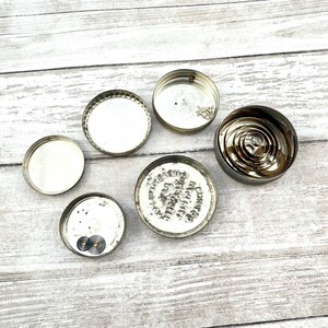 May include: An assortment of small, circular, silver-colored metal objects, including lids and a coiled spring mechanism. Some lids have visible markings and components. The items are arranged on a light-colored, weathered wooden surface.