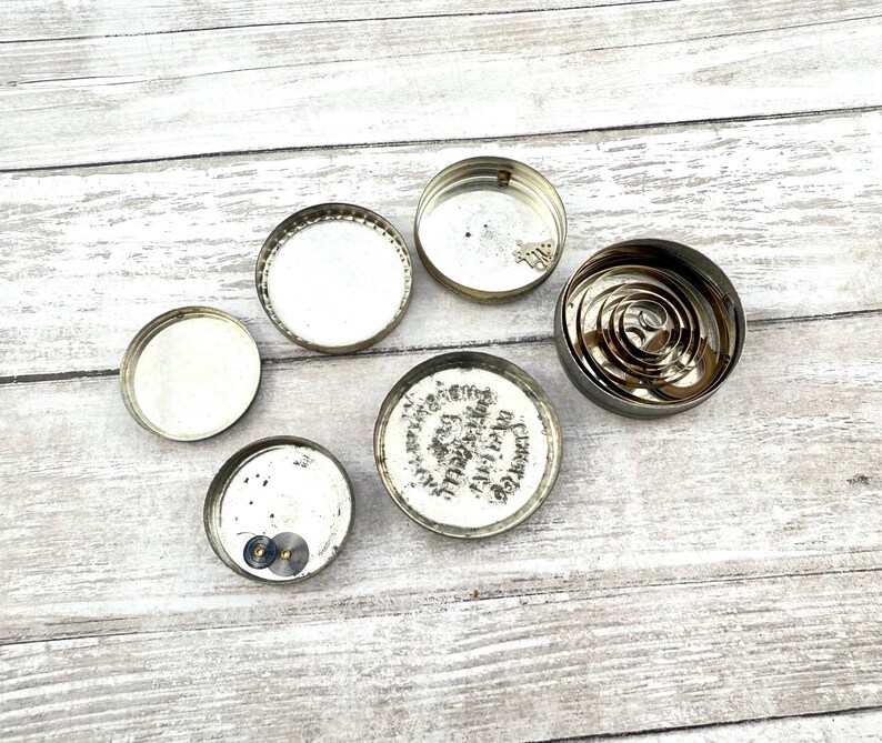 May include: An assortment of small, circular, silver-colored metal objects, including lids and a coiled spring mechanism. Some lids have visible markings and components. The items are arranged on a light-colored, weathered wooden surface.