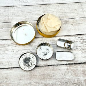 May include: An assortment of small, open metal containers and lids. A yellow tin holds a bundle of tissue paper. Two small rectangular tins and two round lids are also visible. The items are arranged on a light-colored wooden surface.