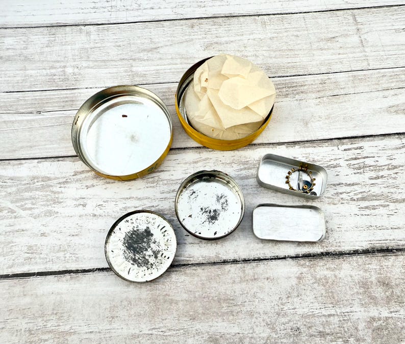 May include: An assortment of small, open metal containers and lids. A yellow tin holds a bundle of tissue paper. Two small rectangular tins and two round lids are also visible. The items are arranged on a light-colored wooden surface.