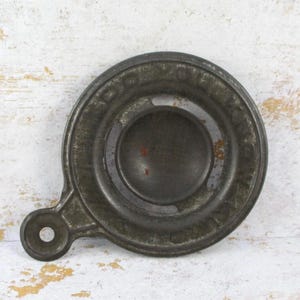 May include: Antique metal object with a circular design, featuring a central disc and an outer ring with embossed text. A small handle with a hole extends from the side. The object has a weathered, aged appearance with signs of rust.