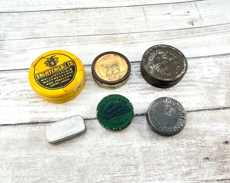 May include: A collection of vintage metal tins in various shapes and sizes. The tins are yellow, green, and silver, with text and logos. One yellow tin reads "SWARTCHILD & CO." and another green tin reads "BENJ ALLEN & CO. Watch Materials CHICAGO".