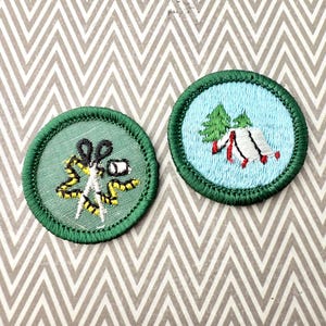May include: Two round embroidered patches with green borders. One features scissors, a measuring tape, and a spool of thread on a green background. The other shows a tent and trees on a light blue background. Both are on a patterned surface.