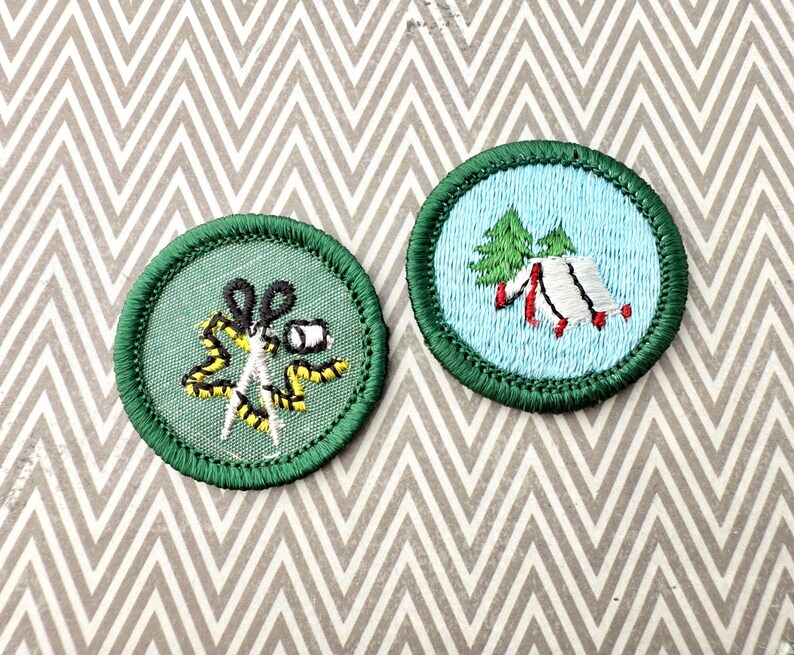 May include: Two round embroidered patches with green borders. One features scissors, a measuring tape, and a spool of thread on a green background. The other shows a tent and trees on a light blue background. Both are on a patterned surface.