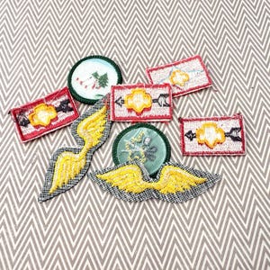 May include: A collection of embroidered patches in various shapes. Includes rectangular patches with yellow and black designs on a red background, circular patches with green borders, and yellow wing-shaped patches. The patches are on a gray and white chevron patterned surface.