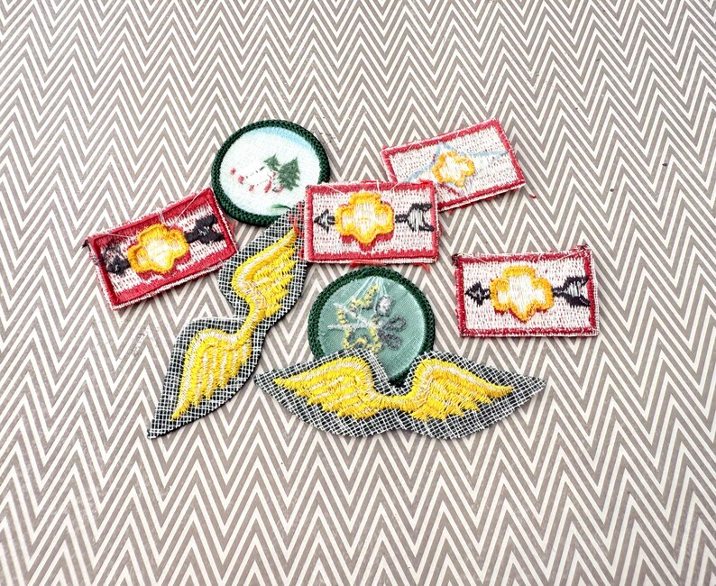 May include: A collection of embroidered patches in various shapes. Includes rectangular patches with yellow and black designs on a red background, circular patches with green borders, and yellow wing-shaped patches. The patches are on a gray and white chevron patterned surface.