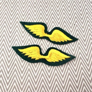 May include: Two embroidered patches in the shape of wings. The wings are yellow with a dark green border. The patches are on a gray and white chevron patterned background.