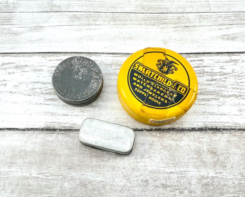 May include: Three vintage metal tins of varying shapes and sizes. A round, dark gray tin with embossed text, a yellow tin with the text "SMARTCHILD & CO.", and a small rectangular tin with text.