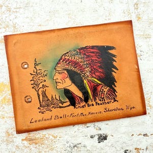 May include: A rectangular tan leather cover featuring a hand-painted illustration of a person in a red and black feathered headdress. The text reads Chief Big Feather, Leeland Ball-Fort MacKenzie, Sheridan, Wyo. Two metal eyelets are visible on the left.