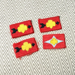 May include: Four red embroidered patches with yellow and black designs. Three patches feature a yellow flower-like shape with black arrows, while the fourth has a yellow flower within a light blue star.