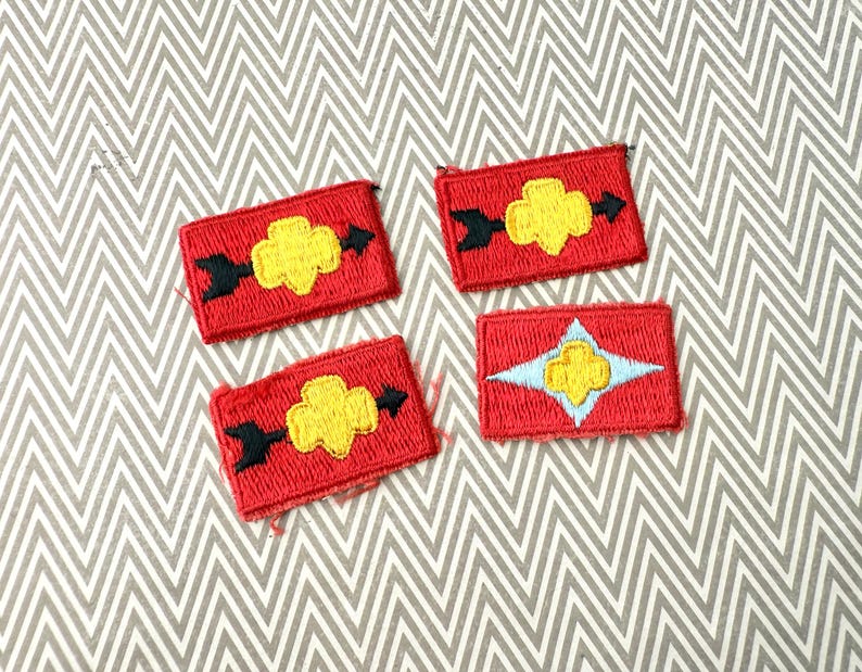 May include: Four red embroidered patches with yellow and black designs. Three patches feature a yellow flower-like shape with black arrows, while the fourth has a yellow flower within a light blue star.