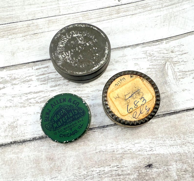 May include: Three vintage metal tins with text. One is dark gray with faded text, another is green with "BENJ. ALLEN & CO Watch Materials and Tools" and the third has a paper label with handwritten numbers.
