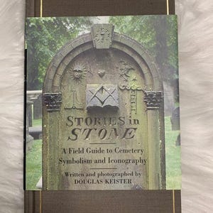 Op de afbeelding: Een hardcover boek getiteld "STORIES in STONE" met een stofomslag met een grafsteen op een begraafplaats. Het boek is bruin met gouden details. De stofomslag luidt "A Field Guide to Cemetery Symbolism and Iconography".