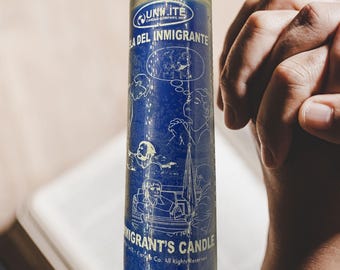 Puede incluir: Una vela de vidrio azul con el texto "LA DEL INMIGRANTE" y "IMMIGRANT'S CANDLE". La vela tiene ilustraciones de personas y bocadillos. La vela está frente a un libro abierto.
