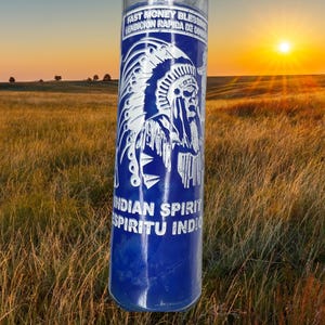 May include: A tall, cylindrical glass candle with a blue label featuring a white Native American chief design. The label includes the text "Fast Money Blessing" and "Indian Spirit". The candle is set against a backdrop of a grassy field and a sunset.