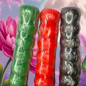 May include: Three vibrantly colored, cylindrical objects are displayed. The objects are green, red, and black, each with a textured, segmented design. They are wrapped in clear plastic, with a soft-focus background featuring a pink flower and a moon.