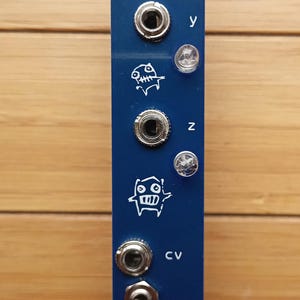 Può includere: Un modulo elettronico blu scuro con il testo "cndl abra" e vari ingressi e uscite. È dotato di una manopola, diversi jack e spie luminose. Il modulo presenta una serie di icone grafiche bianche e il testo "CV".