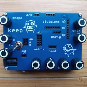 May include: A blue electronic music synthesizer with knobs and a toggle switch. The device has the text "keep" and "a/b" printed on it. There are several knobs with white rings around them. The device has a cartoon monster graphic on it.