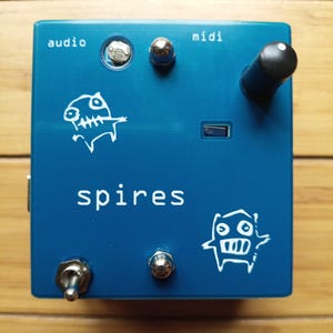 May include: A blue electronic device with the word "spires" in white text. It has various inputs and outputs labeled "audio" and "midi". Two white cartoon fish are printed on the device. A black knob and a silver toggle switch are also visible.