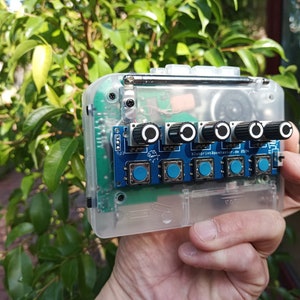 以下が含まれることがあります： 透明なプラスチックケースの中に青い回路基板が入っています。基板には黒いノブが5つと青いボタンが5つ付いています。基板には「BlueprintSpectaclar.de 2014」という文字が印刷されています。