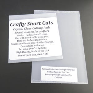 May include: Two clear plastic cutting pads with white labels. The labels read "Crafty Short Cuts" and detail the pads' features, including compatibility with die-cut systems. The pads are 4x4 and 4x6 inches in size. The bottom label warns to remove the protective coating before use.