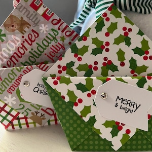 May include: Close-up of three Christmas-themed gift boxes. The boxes feature festive patterns, including holly leaves, polka dots, and holiday phrases. Each box has a white tag with holiday greetings. The boxes are folded paper.